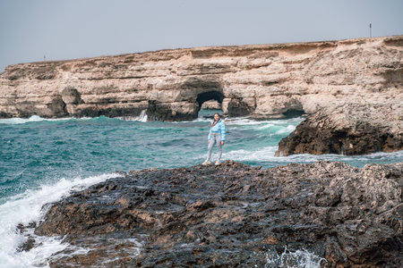 A woman in a blue jacket stands on a rock above a cliff above the sea and looks at the raging ocean. Girl traveler rests, thinks, dreams, enjoys nature. Peace and calm landscape, windy weather.の写真素材
