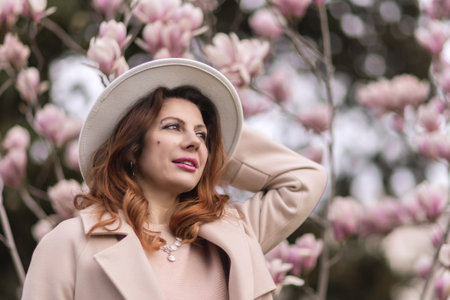 Woman magnolia flowers, surrounded by blossoming trees., hair down, white hat, wearing a light coat....
