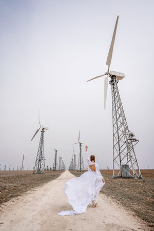 A woman in a white dress is walking down a dirt road in front of a row of wind turbines.の写真素材