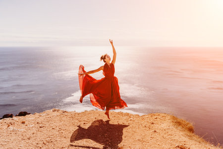 Woman red dress sea. Female dancer in a long red dress posing on a beach with rocks on sunny day. Girl on the nature on blue sky background.の写真素材