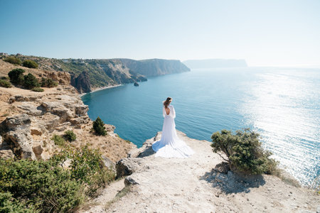 woman white dress stands on a rocky cliff overlooking the ocean. The scene is serene and peaceful, with the womans dress billowing in the wind.の写真素材