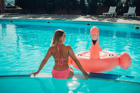 A woman in a pink bikini is sitting on a pink inflatable flamingo in a pool. The scene is playful...