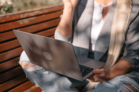A woman is sitting on a bench with a laptop in front of her. She is wearing a gray jacket and a white shirt.の写真素材