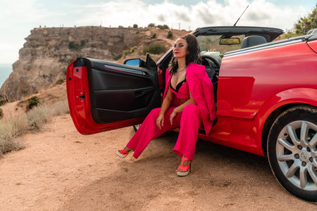 A woman in a pink suit sits in the open door of a red convertible. The scene is set on a rocky hillside overlooking the ocean. The woman is posing for a photo.の写真素材