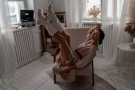 A woman is sitting in a chair with her legs spread apart. She is wearing white boots and a white dress. The room is decorated with a white color theme.の写真素材