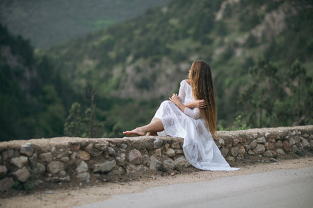 A woman in a white dress is sitting on a stone wall overlooking a mountain. The scene is serene and peaceful, with the womans long hair blowing in the wind. Concept of calm and tranquility.の写真素材