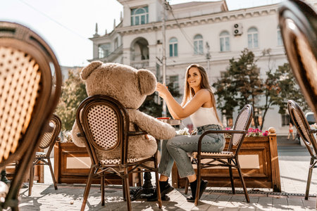 A woman sits cafe with a teddy bear next to her. The scene is set in a city with several chairs and tables around her. The woman is enjoying her time at the outdoor cafe.の写真素材