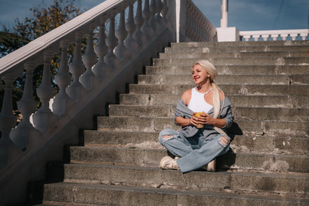 A woman sits on a set of stairs, holding a cup of coffee. She is enjoying her coffee and taking in the view from the top of the stairs.の写真素材