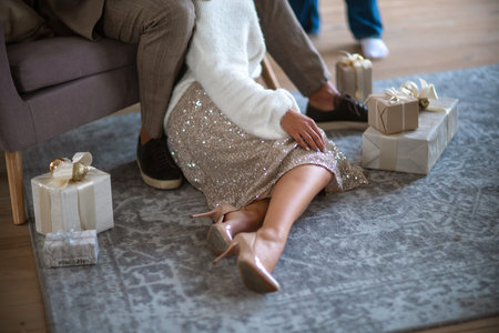 A romantic couple, a man sitting on the sofa and a woman on the carpet next to surrounded by gifts on a gray carpet. She is wearing a light skirt and beige high-heeled shoes.の写真素材