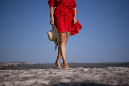 A woman in a red dress is standing on a rocky beach with a straw hat in her handの写真素材