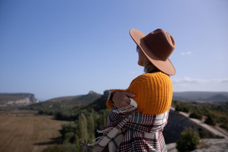 A woman wearing a brown hat and a yellow sweater is standing on a hillsideの写真素材