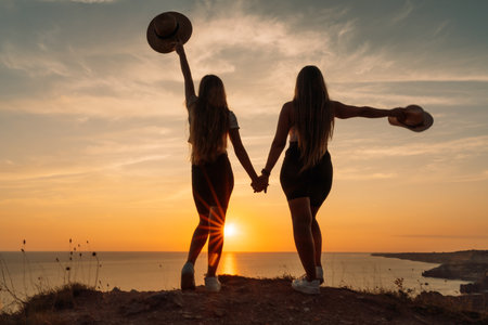 Two women are standing on a hillside, holding hands and wearing hats. The sun is setting in the background, creating a warm and peaceful atmosphere.の写真素材