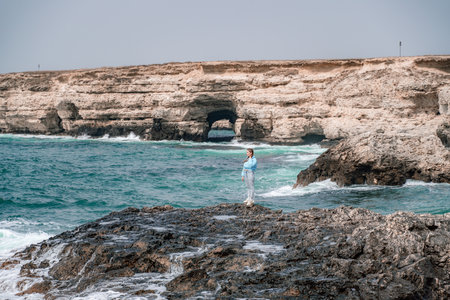 A woman in a blue jacket stands on a rock above a cliff above the sea and looks at the raging ocean. Girl traveler rests, thinks, dreams, enjoys nature. Peace and calm landscape, windy weather.の写真素材