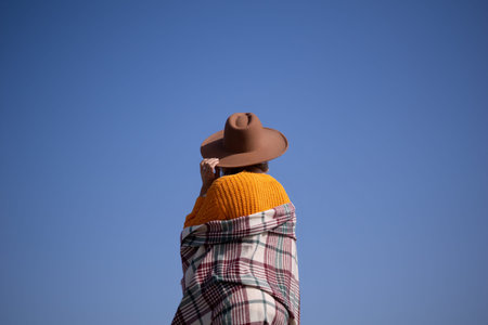 A woman wearing a brown hat and a plaid blanket stands in front of a blue skyの写真素材