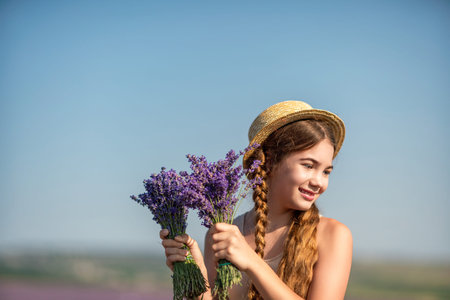 girl is holding a bunch of lavender purple flowers in her hands and wearing a straw hat. She is smiling and she is enjoying herself. The scene is set in a field of lavender, which adds to the peaceful.の写真素材