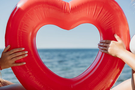 Two women are holding a red inflatable heart on the beach