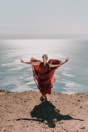 Woman red dress sea. Female dancer posing on a rocky outcrop high above the sea. Girl on the nature on blue sky background. Fashion photo.の写真素材