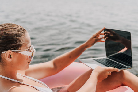 A woman is sitting in a pink inflatable raft with a laptop on her lap. She is wearing sunglasses and...