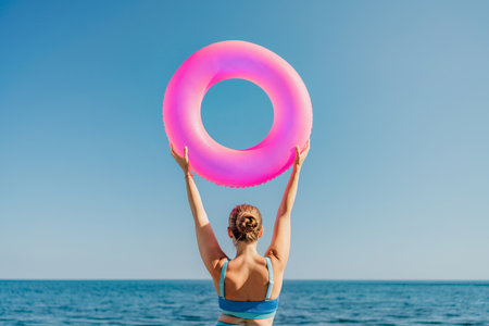A woman is holding a pink inflatable ring above the ocean. Concept of fun and relaxation, as the woman is enjoying her time at the beach. The pink color of the ring adds a playful.の写真素材