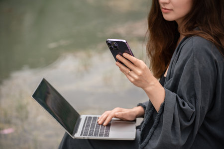 A woman is sitting on the ground with a laptop and a cell phone in her hands. She is using the laptop to browse the internet and the cell phone to communicate with someone.の写真素材