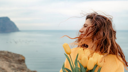 Portrait of a happy woman with hair flying in the wind against the backdrop of mountains and sea. Holding a bouquet of yellow tulips in her hands, wearing a yellow sweaterの写真素材