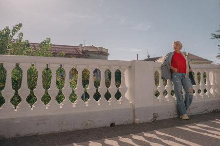 A woman in a gray jacket and blue jeans is leaning against a white railing. She is holding a book in...