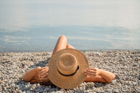 A woman is laying on the beach wearing a straw hat. She is relaxed and enjoying the sun.の写真素材