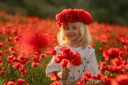 A young girl is standing in a field of red flowers, wearing a red flower crown. She is smiling and holding a bouquet of red flowers. Concept of joy and innocence.の写真素材