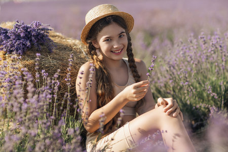 A young girl is sitting in a field of purple flowers, holding a purple flower in her hand. She is smiling and she is enjoying the moment. Concept of happiness and contentment.の写真素材