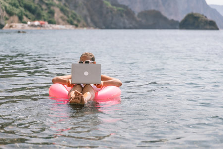 A woman is floating in a pink inflatable raft with a laptop in her hand. She is smiling and she is enjoying her time in the water.の写真素材