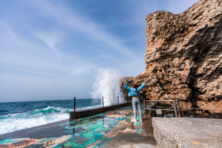 A woman in a blue jacket stands on a rock above a cliff above the sea and looks at the raging ocean. Girl traveler rests, thinks, dreams, enjoys nature. Peace and calm landscape, windy weather.の写真素材