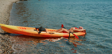 A woman is laying on a kayak in the water. The kayak is orange and has a paddle on it. The woman is wearing a red hat.の写真素材