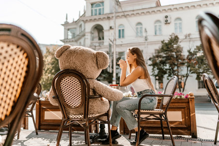 A woman sits cafe with a teddy bear next to her. The scene is set in a city with several chairs and tables around her. The woman is enjoying her time at the outdoor cafe.の写真素材