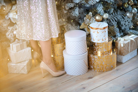 Womens legs. Cropped close-up photo of healthy beautiful elegant female legs in beige high-heeled shoes, a woman stands on the background of a Christmas tree with giftsの写真素材