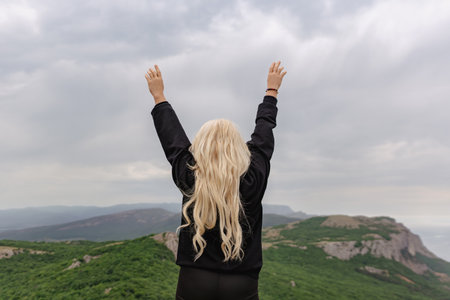 A woman with blonde hair is standing on a mountain, looking up at the skyの写真素材