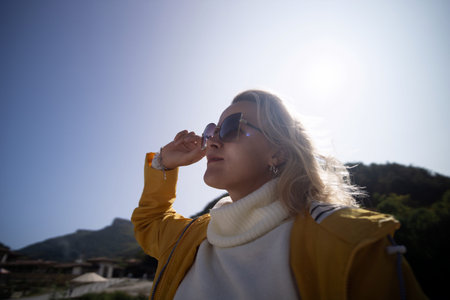 A woman wearing a yellow jacket and sunglasses is standing in the sun