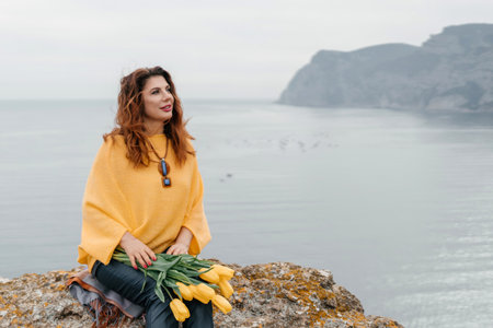 A woman in a yellow sweater sits on a rock by the ocean, holding a bouquet of yellow flowers. Concept of tranquility and relaxation, as the woman enjoys the peaceful view of the water.の写真素材