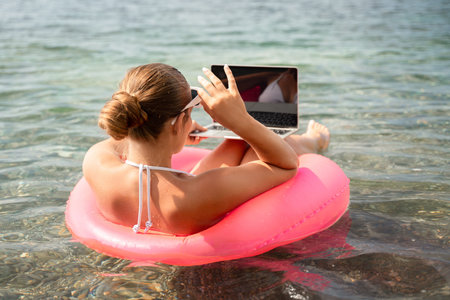 A woman is sitting on a red inflatable raft with a laptop on her lap. She is wearing a white bikini and sunglasses. Concept of relaxation and leisure.の写真素材