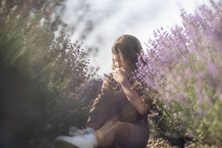 Girl is sitting in a field of purple flowers. She is holding a basket of flowers and smiling. Scene is peaceful and serene, as the girl is surrounded by the beauty of nature.の写真素材