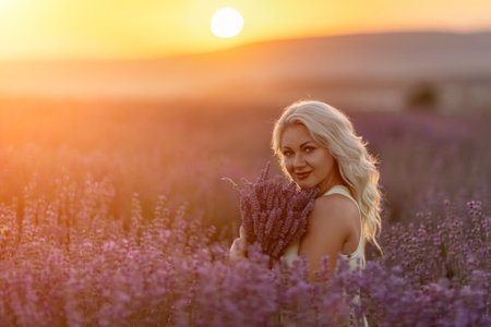 Blonde woman poses in lavender field at sunset. Happy woman in white dress holds lavender bouquet. Aromatherapy concept, lavender oil, photo session in lavenderの写真素材