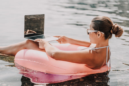 A woman is sitting in a pink inflatable raft on a lake, using a laptop. Concept of relaxation and leisure, as the woman is enjoying her time outdoors while working on her laptop.の写真素材