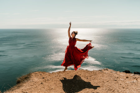 Woman red dress sea. Female dancer posing on a rocky outcrop high above the sea. Girl on the nature on blue sky background. Fashion photo.の写真素材