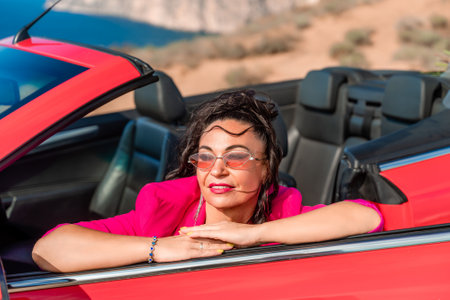A woman in a pink jacket is sitting in a red convertible. She is wearing sunglasses and has her hand on her hip. Scene is relaxed and carefree.の写真素材