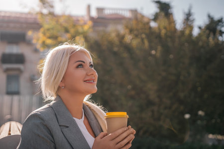A blonde woman is sitting on a bench and holding a cup of coffee. She is smiling and looking up at the sky.の写真素材
