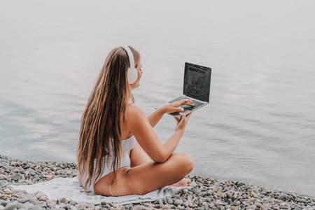 A woman is sitting on the beach with a laptop open in front of her. She is wearing a white bikini and has long hair. Concept of relaxation and leisure.の写真素材