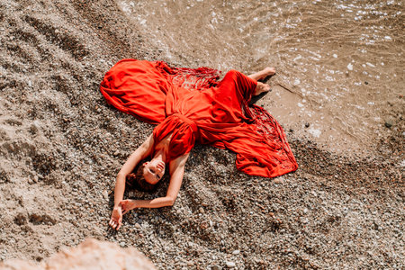 Woman red dress sea. Female dancer in a long red dress posing on a beach with rocks on sunny dayの写真素材