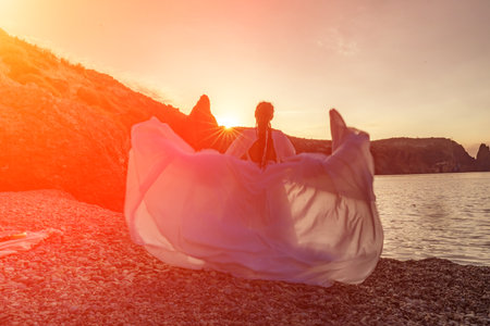 Mysterious woman silhouette long hair walks on the beach ocean water, sea nymph wind listens to the wave. Throws up a long white dress, a divine sunset. Artistic photo from the back without a faceの写真素材