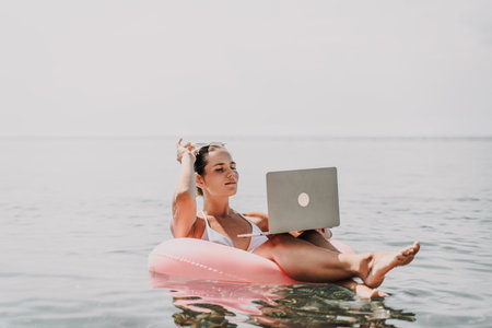 A woman is sitting on a pink inflatable raft in the ocean, using a laptop. Concept of relaxation and...