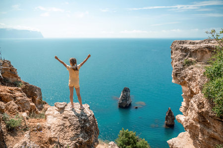 Happy girl stands on a rock high above the sea, wearing a yellow jumpsuit and sporting braided hair, depicting the idea of a summer vacation by the sea.の写真素材
