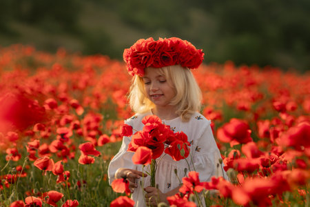 A young girl is standing in a field of red poppies. She is wearing a red hat and holding a bouquet of red flowers. The scene is peaceful and serene, with the girl surrounded by the beauty of nature.の写真素材